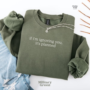 May include: Military green sweatshirt with the phrase "if I'm ignoring you, it's planned" in white lettering. A silver necklace with a pendant is draped across the front. The sweatshirt is folded with the sleeves cuffed.