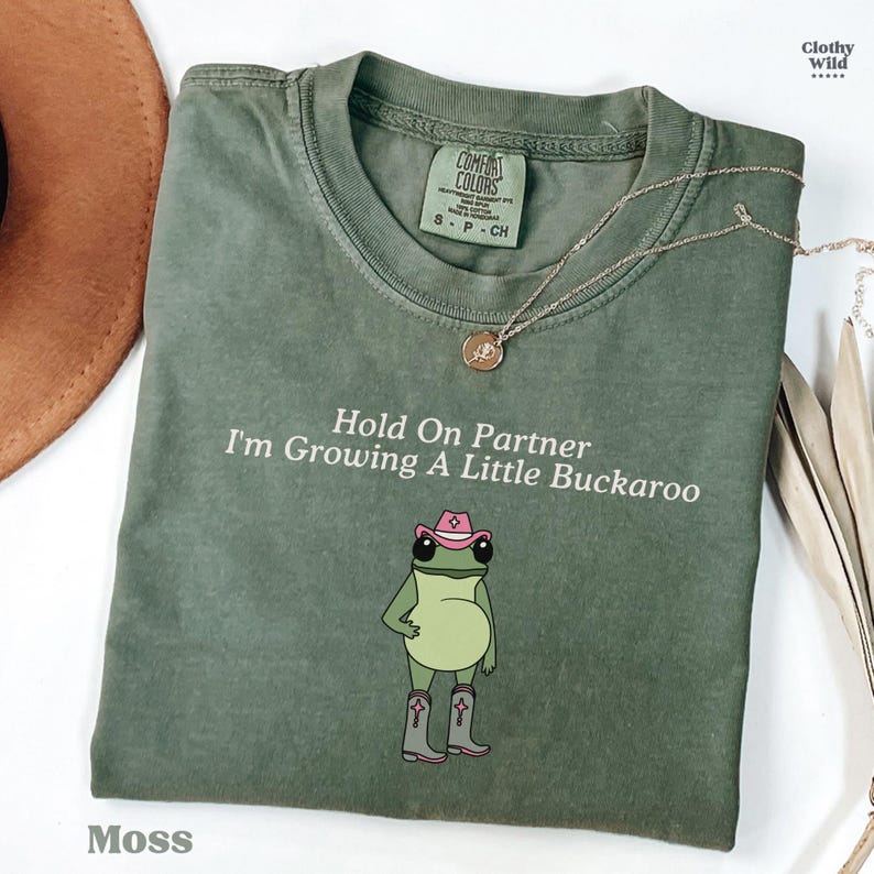 May include: Moss green t-shirt with the text "Hold On Partner I'm Growing A Little Buckaroo" and a cartoon frog wearing a pink cowboy hat and boots. A gold necklace is draped across the shirt.