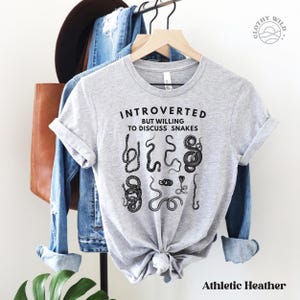 Puede incluir: Una camiseta gris con el texto "Introvertido pero dispuesto a hablar de serpientes" y un gr&aacute;fico de varias serpientes. La camiseta est&aacute; colgada de una percha.