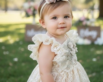 Könnte beinhalten: Cremefarbenes Spitzenkleid mit Rüschenärmeln und passendem Stirnband. Das Stirnband hat das Wort "one" in glitzernden Verzierungen. Das Kleid hat detaillierte Spitze und einen floralen Akzent.