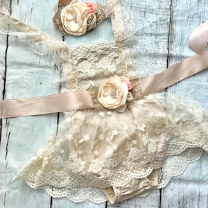 May include: A beige lace baby dress with a satin ribbon and a flower headband. The dress has a scalloped lace trim and a ruffled skirt. The headband has a large flower with a rhinestone center.