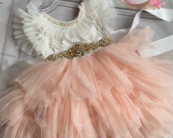 May include: A white and peach tulle dress with a lace bodice and a gold beaded belt. The dress has a ruffled neckline and a layered skirt.