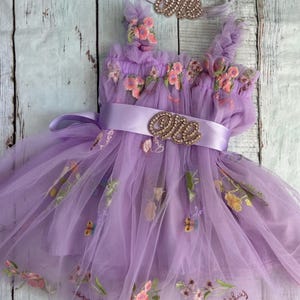 Puede incluir: Un vestido de tul lavanda con bordado floral y una diadema a juego. El vestido tiene una faja de satén con un adorno de "uno" dorado. La diadema también tiene un adorno de "uno" dorado.