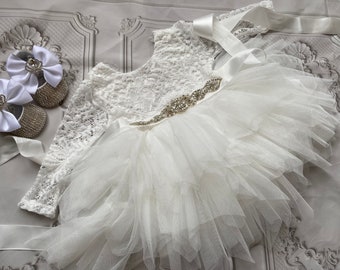 May include: White lace baby dress with a tulle skirt and a rhinestone belt. The dress has long sleeves and a round neckline. The dress is paired with white satin shoes with rhinestone embellishments.