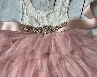 May include: A blush pink tulle dress with a white lace bodice and a rose gold rhinestone belt. The dress also includes a matching rose gold rhinestone headband.