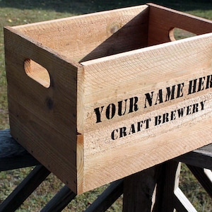 May include: A wooden crate with the text "YOUR NAME HERE CRAFT BREWERY" printed in black. The crate is made of light brown wood and has cut-out handles on the sides. It is sitting on a wooden railing.