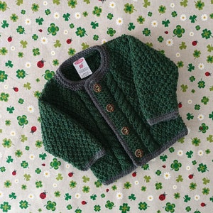 Könnte beinhalten: Ein grüner gestrickter Baby-Cardigan mit Zopfmuster und grauem Besatz. Der Cardigan hat drei braune Knöpfe auf der Vorderseite.
