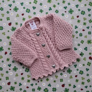 Könnte beinhalten: Ein rosa gestrickter Baby-Cardigan mit Zopfmuster und herzförmigen Knöpfen. Der Cardigan hat einen gewellten Saum und ist aus weichem Garn gefertigt.
