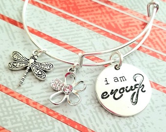 May include: Silver bangle bracelet with three charms: a dragonfly, a flower with rhinestones, and a round charm with the text "i am enough".