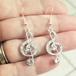 May include: Silver-coloured treble clef earrings. Each earring has a treble clef shape, studded with small, clear crystals. Larger, round crystals are at the centre and bottom of each clef, with a fish hook style ear wire.