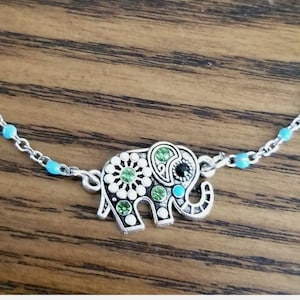 Op de afbeelding: Een zilveren ketting met een turquoise blauwe kralenaccent en een zilveren olifantbedel. De olifantbedel is versierd met groene en witte strass.
