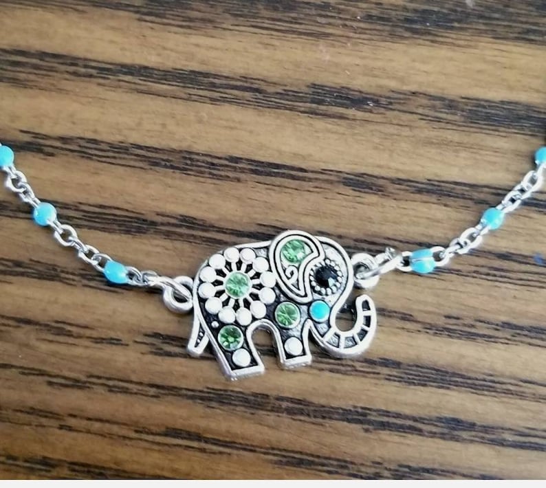 Op de afbeelding: Een zilveren ketting met een turquoise blauwe kralenaccent en een zilveren olifantbedel. De olifantbedel is versierd met groene en witte strass.