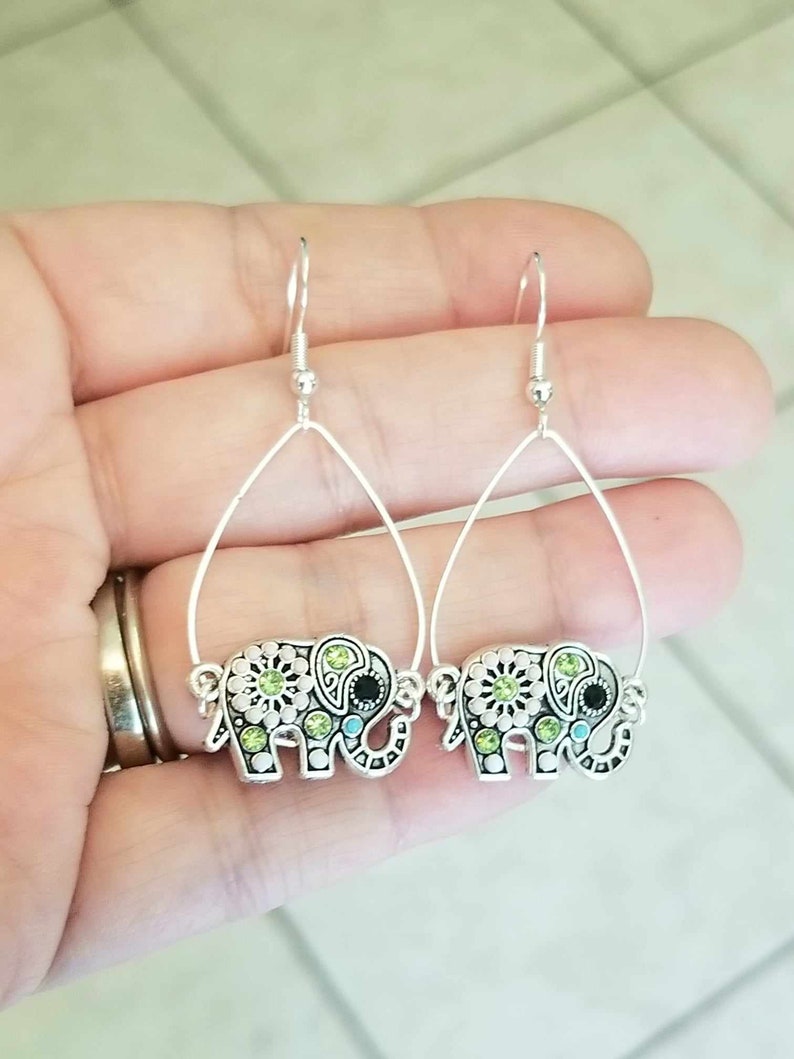Op de afbeelding: Een paar zilveren oorhangers met een olifantendesign met groene en blauwe strass.