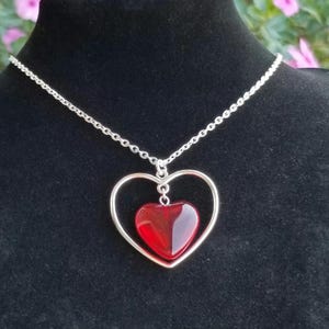 May include: A silver-toned necklace featuring a heart-shaped pendant. The pendant has a larger, open heart frame with a smaller, solid red heart suspended inside. The chain is delicate and the background is black.