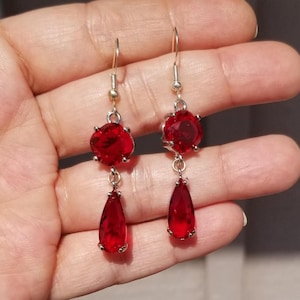 May include: A pair of dangle earrings featuring bright red, faceted gemstones. Each earring has a round top stone and a teardrop-shaped bottom stone, all set in silver-colored metal. The earrings are suspended from silver-colored ear wires.