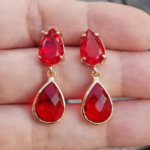 May include: A pair of gold-toned earrings featuring two faceted, teardrop-shaped red gemstones. The earrings have a classic design with a smaller gem at the top and a larger one dangling below. The gems are a vibrant, rich red color.