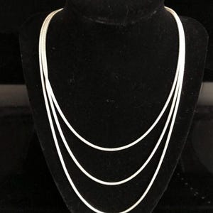 May include: A silver necklace with three strands of varying lengths. The necklace is displayed on a black velvet bust. The strands are smooth and reflective, creating a simple yet elegant design.