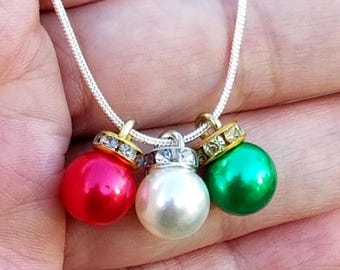 Puede incluir: Un collar plateado con tres colgantes de adorno navideño. Los adornos son rojos, blancos y verdes, cada uno con una tapa dorada adornada con cristales transparentes. Un accesorio festivo.