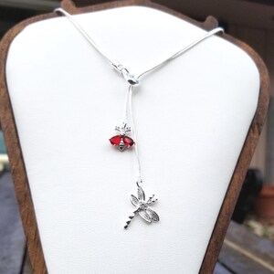 Puede incluir: Un collar plateado con un colgante de libélula y una gema roja. El colgante de libélula está adornado con cristales transparentes. El collar se exhibe sobre un busto blanco contra un fondo exterior borroso.