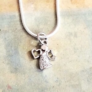 Puede incluir: Un collar de cadena de plata con un peque&ntilde;o colgante de &aacute;ngel brillante. El &aacute;ngel tiene un halo y alas en forma de coraz&oacute;n.