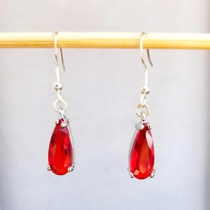 May include: A pair of drop earrings featuring faceted, pear-shaped red stones set in silver-toned metal. Each earring hangs from a simple silver-toned hook wire, showcasing a vibrant crimson hue and a polished, reflective finish on the stones.
