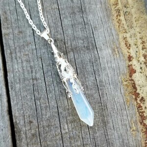 May include: A silver pendant necklace featuring a light blue and white crystal pendant. The crystal is encased in a decorative silver filigree setting and hangs from a delicate silver chain. The pendant is approximately 2 inches long.