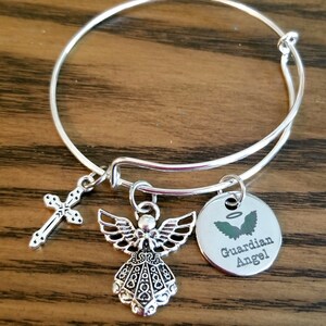 May include: Silver bangle bracelet with a charm of an angel, a cross, and a round charm with the words "Guardian Angel" and a halo above a pair of wings.