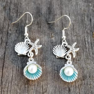 May include: A pair of silver-toned dangle earrings with a beach theme. Each earring features a seashell, starfish, and a shell with a pearl. The shell has a turquoise interior. The earrings hang from a silver hook.