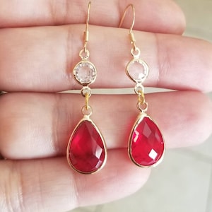 May include: Gold dangle earrings with a teardrop-shaped red gemstone and a smaller clear round gemstone.