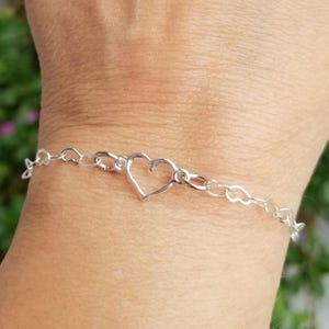 May include: A delicate silver bracelet featuring a heart-shaped charm as the focal point. The bracelet is composed of small, interconnected links, with a larger heart-shaped pendant at the center. The design is simple and elegant, suitable for everyday wear.