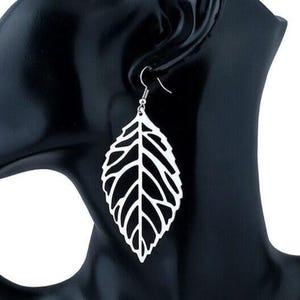 May include: A silver-colored leaf-shaped earring with intricate vein detailing. The earring hangs from a simple hook, showcasing a delicate, openwork design. The earring is displayed against a black background.