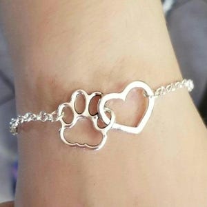 Peut inclure: Un bracelet en argent avec un charme en forme de patte et un charme en forme de cœur. Le bracelet est porté au poignet d'une personne.