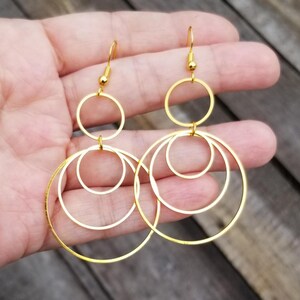 May include: A pair of gold-toned dangle earrings featuring a multi-layered design of interconnected circular hoops. Each earring consists of three thin, polished gold rings of varying sizes suspended from a standard fishhook-style ear wire.