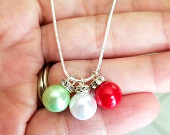 Puede incluir: Un collar de cadena de serpiente plateada con tres cuentas redondas y lisas en verde claro, blanco y rojo. Cada cuenta está rematada con una pequeña tapa plateada incrustada con cristales claros y brillantes, creando un colgante festivo.
