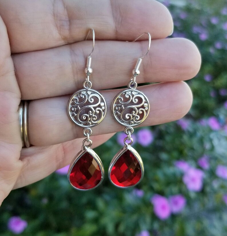 以下が含まれることがあります： 赤い雫型のクリスタルが付いたシルバーのドロップイヤリングのペア。イヤリングは、円形のトップとぶら下がっているクリスタルを持つフィリグリーのデザインが特徴です。