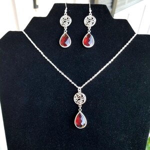 以下が含まれることがあります： 赤い雫型の石のペンダントとそれに合わせたイヤリングが付いた、シルバーのネックレスとイヤリングのセット。ペンダントとイヤリングは繊細なフィリグリーのデザインが施されています。