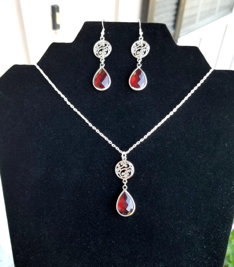 以下が含まれることがあります： 赤い雫型の石のペンダントとそれに合わせたイヤリングが付いた、シルバーのネックレスとイヤリングのセット。ペンダントとイヤリングは繊細なフィリグリーのデザインが施されています。