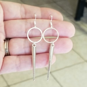 Puede incluir: Pendientes de plata con una gota larga y puntiaguda y un pequeño círculo en la parte superior.