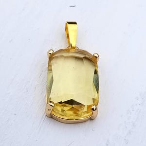 Könnte beinhalten: Ein rechteckiger Anhänger mit einem facettierten, durchscheinenden gelben Edelstein, der in einer polierten goldfarbenen Metallfassung mit vier Krappen sitzt und oben eine passende goldfarbene Öse zur Befestigung an einer Kette besitzt.