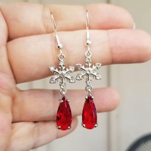 Könnte beinhalten: Silberne Schneeflocken-Ohrringe mit roten, tropfenförmigen Edelsteinen. Die Ohrringe haben einen Hakenverschluss. Das Schneeflocken-Design ist mit kleinen, klaren Kristallen versehen. Festlicher Schmuck für den Winter.