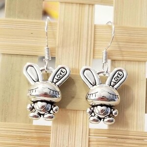 May include: A pair of silver-toned metal drop earrings featuring a stylized rabbit character with closed eyes and a heart on its chest. One ear of each rabbit is engraved with the word LOVE. The earrings hang from simple silver-toned metal hooks.