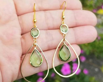 May include: Gold dangle earrings with two green gemstone drops. The earrings have a teardrop shape and are suspended from a small gold hoop.