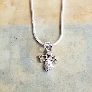 Puede incluir: Un collar de cadena de plata con un peque&ntilde;o colgante de &aacute;ngel brillante. El &aacute;ngel tiene alas y un halo en forma de coraz&oacute;n.