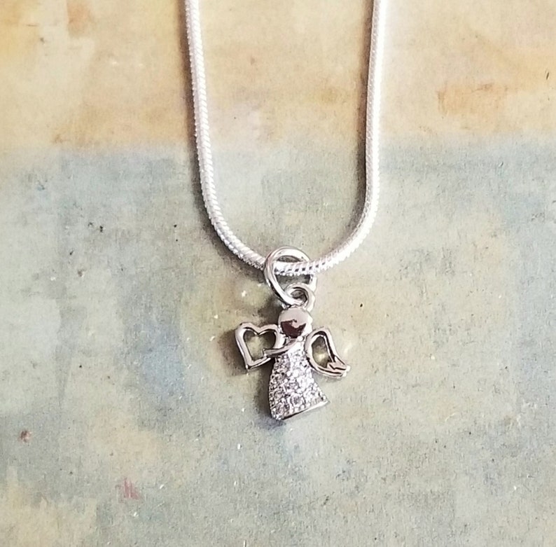 Puede incluir: Un collar de cadena de plata con un peque&ntilde;o colgante de &aacute;ngel brillante. El &aacute;ngel tiene alas y un halo en forma de coraz&oacute;n.
