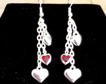 May include: Silver dangle earrings with red heart charms. The earrings have multiple chains and hearts.