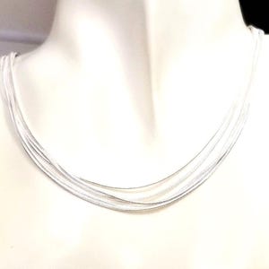 May include: A multi-strand silver necklace. The necklace features several delicate strands of silver, creating a layered effect. The strands drape gracefully, showcasing a simple yet elegant design. The jewelry is displayed on a white background.