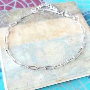 May include: A silver chain bracelet with a delicate, elongated link design. The bracelet is displayed on a light blue and beige surface, showcasing its polished, reflective surface. The bracelet is a jewelry item.