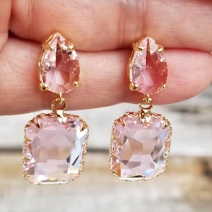 May include: A pair of gold-toned drop earrings featuring faceted light pink crystals. Each earring consists of a smaller pear-shaped crystal at the top connected by a jump ring to a larger, rectangular-shaped crystal base with scalloped edges.