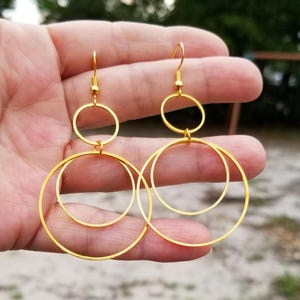 May include: A pair of gold-toned dangle earrings featuring a tiered design. Each earring consists of two interconnected circular hoops of varying sizes, suspended from a simple hook wire, creating a layered, geometric, and metallic appearance.