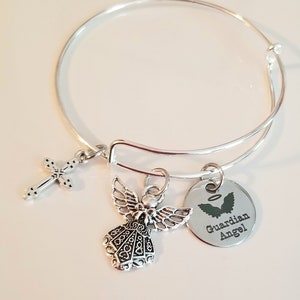 May include: Silver bangle bracelet with a charm of an angel, a cross, and a round charm with the text "Guardian Angel".